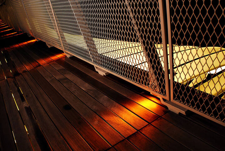 Close detail of warm linear light grazing timber decking beneath the mesh balustrade